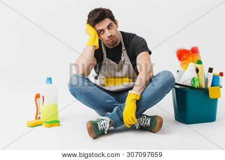 Image of a tired bored young houseman househusband with cleansers on floor isolated over white wall background.