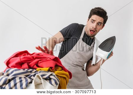 Image of a young scared confused houseman househusband holding iron looking at clothes isolated over white wall background.
