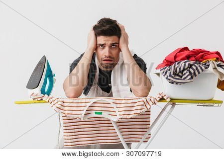 Image of a tired bored young houseman househusband with iron and clothes isolated over white wall background.