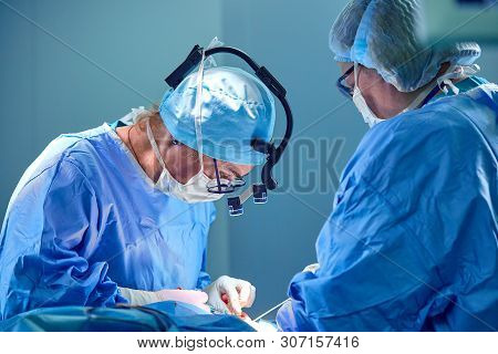 Surgeon And His Assistant Performing Cosmetic Surgery In Hospital Operating Room. Surgeon In Mask We