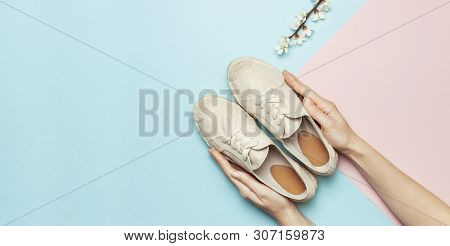 Female Hands Hold Beige Fashionable Espadrilles With Spring Flowering Branch On Pastel Blue Pink Bac