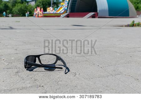 Broken Sun Glasses Lie On A Stone Tiled Walkway In The City Park Against The Background Of Residenti