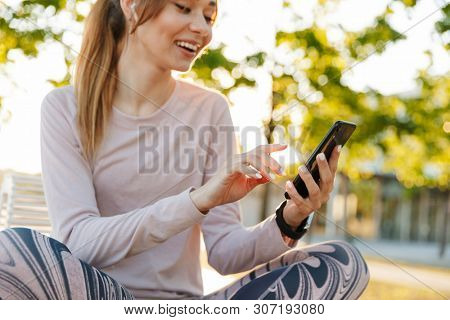 Cropped image of a cheerful young fitness woman relaxing outdoos, sitting on a bench, using mobile phone