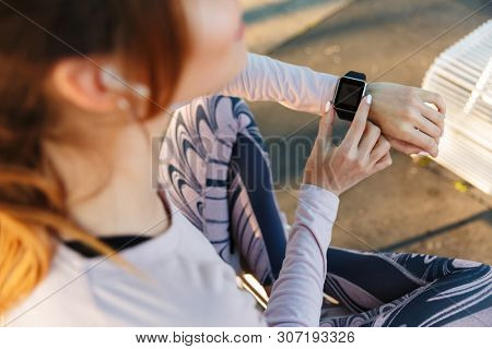Top view close up of a young fitness woman relaxing outdoos, sitting on a bench, using smart watch