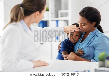 medicine, healthcare and pediatry concept - african american mother with sick son and caucasian doctor measuring baby's temperature by hand at clinic