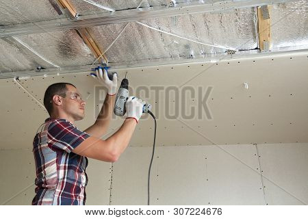Young Man In Goggles Fixing Drywall Suspended Ceiling To Metal Frame Using Electrical Screwdriver On