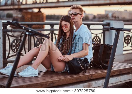 Young Beautiful Romantic Couple With Electric Scooters Is Sitting On The Bench Near Brige At Sunset 
