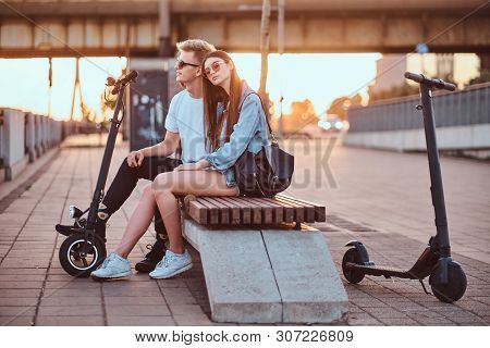 Young Beautiful Romantic Couple With Electric Scooters Is Sitting On The Bench Near Brige At Sunset 