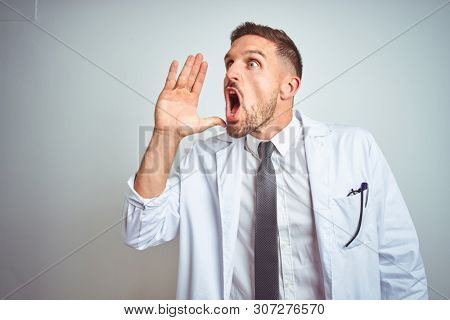 Young handsome doctor man wearing white profressional coat over isolated background shouting and screaming loud to side with hand on mouth. Communication concept.