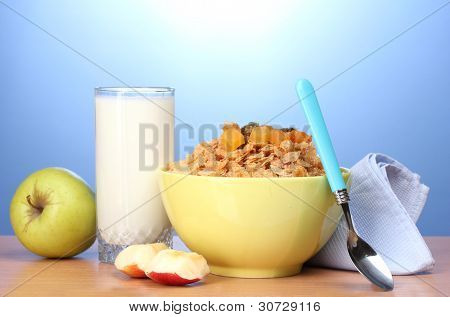 leckere Cornflakes in grüner Schale, Äpfel und Glas Milch auf Holztisch auf blauem Hintergrund