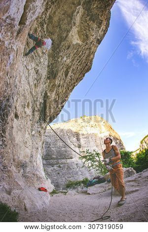 Athletic Man Climbs An Overhanging Rock 8c-9a Category With Rope With Man Belayed Him. Sport Climbin