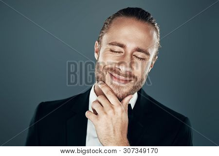 Happy About... Charming Young Man In Bow Tie Smiling And Keeping Hand On Chin While Standing Against