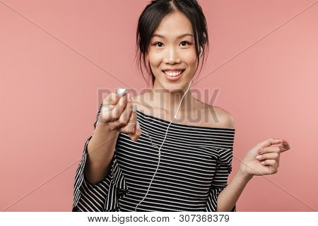 Photo of optimistic asian woman dressed in basic wear holding earphone while listening to music isolated over red background in studio