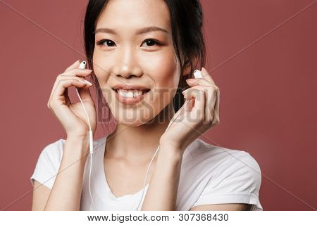 Portrait of optimistic asian woman dressed in basic wear smiling and listening to music with earphones isolated over red background in studio