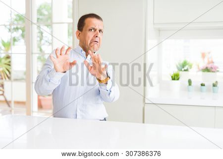 Middle age man sitting at home afraid and terrified with fear expression stop gesture with hands, shouting in shock. Panic concept.