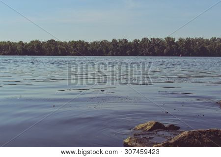 The river landscape with waves on the water surface. The morning tranquility along the River. The forest on the other coast. A blue sky over stream. A few stones in the water.