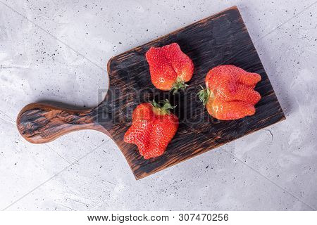 Three Ugly Ripe Red Strawberries On Burned Kitchen Cutting Board On Grey Concrete Background.