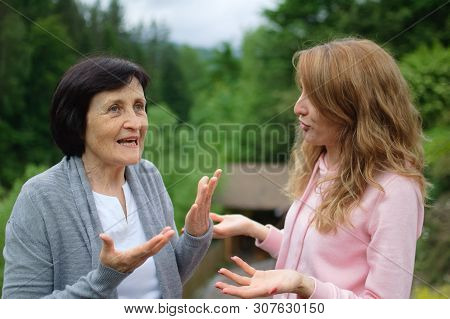 Beautiful Women, Two Generations, Mother And Daughter Are Communicating Outside In Front Of A Blurre