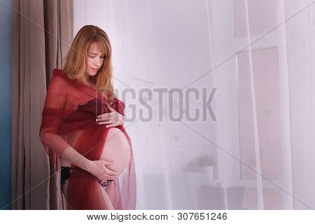 Expectant Mother Stands At The Bedroom Window, Gently Touching The Belly, Which Is Her Future Child