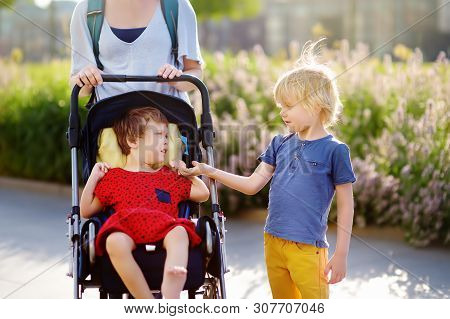 Woman With A Boy And A Disabled Girl In A Wheelchair Walking In The Park Summer. Child Cerebral Pals