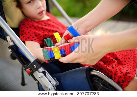 Mother Puts Orthosis On Her Daughter Arms. Disabled Girl Sitting On A Wheelchair. Child Cerebral Pal