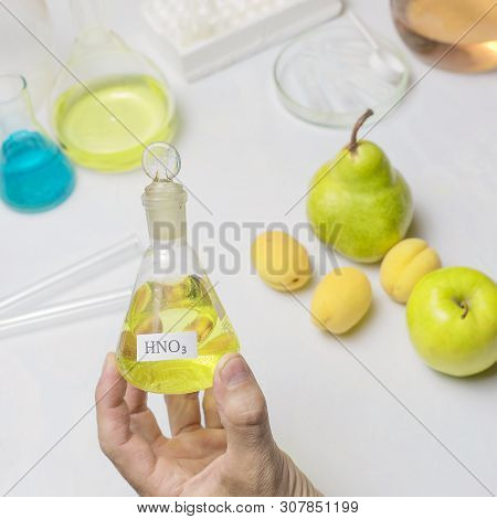The Lab Assistent Hand Holds A Flask Of Nitric Acid On The Background Of The Fruit Lying On The Tabl