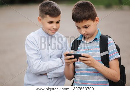 Two Boy Sitting On The Bench And Play Online Games. One Boys With Backpack. Young Boys Use Their Pho