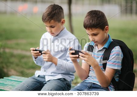 Two Boy Sitting On The Bench And Play Online Games. One Boys With Backpack. Young Boys Use Their Pho