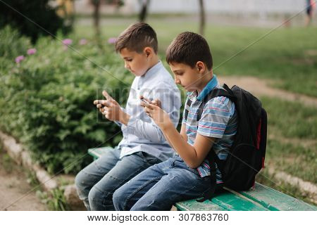 Two Boy Sitting On The Bench And Play Online Games. One Boys With Backpack. Young Boys Use Their Pho