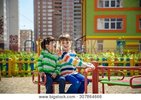 Two Cheerful Boys In T-shirts Play On A Big Iron Carousel On The Playground At The Summer Or Spring