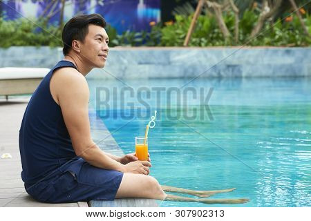 Pensive Mature Vietnamese Man Sitting At Swimming Pool And Drinking Fruit Cocktail