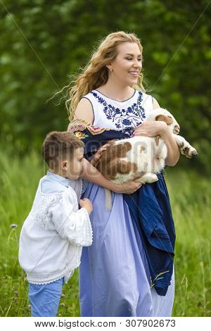 Portrait Of Smiling Mother With Her Little Son. Holding Cub Of Newborn Lamb Outdoors On Nature Backg
