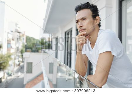 Pensive Vietnamese Man Rubbing Chin And Looking At City Streets When Standing On Bacony Of Hotel Roo