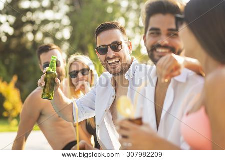 Group Of Friends At A Poolside Summer Party, Sitting At The Edge Of A Swimming Pool, Drinking Cockta