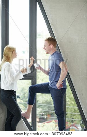 Boyfriend And Girlfriend Standing At Assymetric Big Window, Drinking Coffee And Discussing Plans For