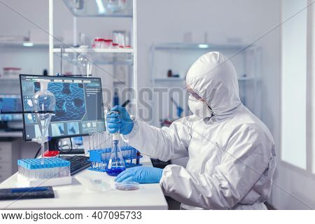 Man Taking Sample Of Liquid From Glass Flask Using Micropipette Dressed In Protection Suit. Chemist 