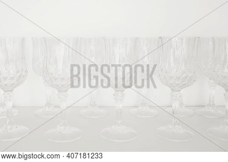 Dishes In Kitchen. Transparent Glass Wineglasses, Stand In Row On White Shelf In Kitchen Cabinet.