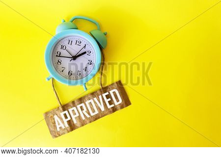 On A Yellow Background There Is An Alarm Clock With A Tag On Which The Text Approved Is Written