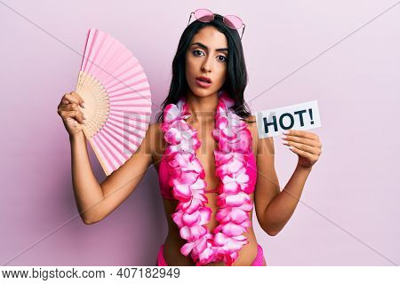 Beautiful hispanic woman wearing bikini and handfan holding hot word in shock face, looking skeptical and sarcastic, surprised with open mouth 