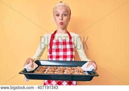 Young blonde woman wearing baker uniform holding homemade cookies puffing cheeks with funny face. mouth inflated with air, catching air. 