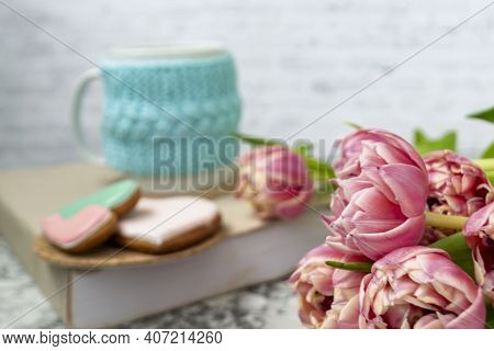 A White Cup Of Tea In A Knitted Blue Case Stands On A Book And Pink Flowers, Tulips And Gingerbread 