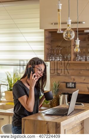 Woman Having A Phone Conversation While Working Remotely From Home; Female Web Developer Using Lapto