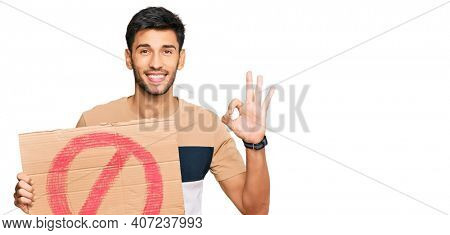 Young handsome man holding prohibited warning banner doing ok sign with fingers, smiling friendly gesturing excellent symbol 
