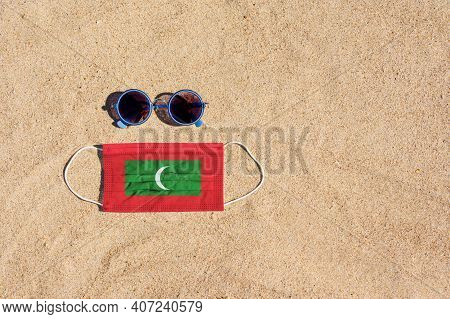 A Medical Mask In The Color Of The Maldives Flag Lies On The Sandy Beach Next To The Glasses. The Co