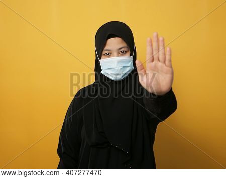 Asian Muslim Businesswoman Wearing Hijab And Medical Face Mask Shows Stop Hand Sign Gesture, Against