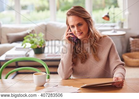 Smiling Woman On Phone Call Sitting At Table At Home With Digital Tablet Reviewing Domestic Finances