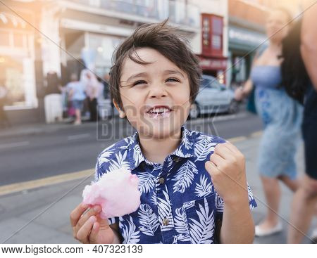 Smiling Kid Eating Cotton Candy With Blurry People Walking On Street Background,young Boy Traveler W