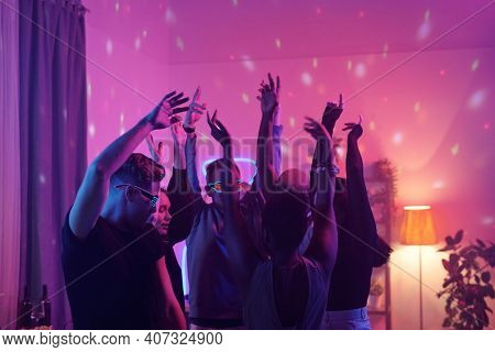 Young intercultural friends in smart casualwear raising arms while dancing together at home party in living-room illuminated with pink lighting