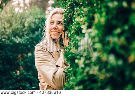 Portrait Of Blonde Smiling Woman In Green Forestor Park In Summer Wearing Trendy Beige Trench Coat