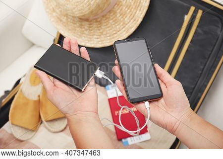 Woman Charging Smartphone With Power Bank Near Packed Suitcase Indoors, Closeup